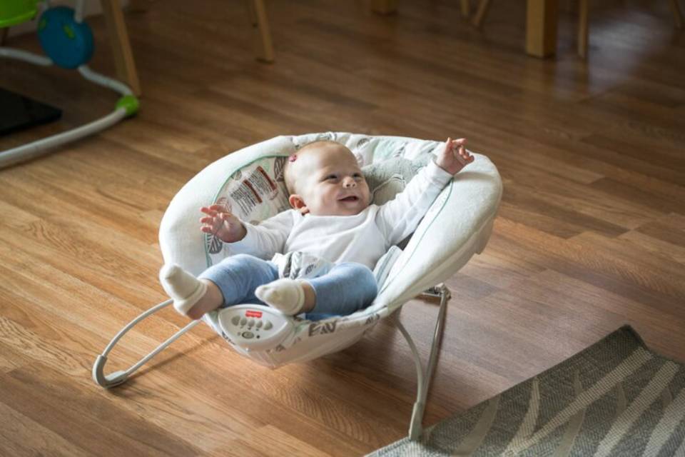 An infant in a cozy bouncer, wearing a white top and blue jeans, stretches out with arms raised, surrounded by a warm wooden floor.