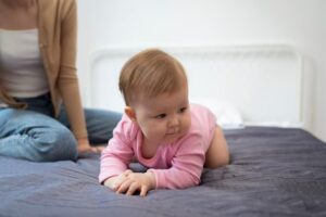 A baby in a pink onesie is crawling on a dark bedspread, with an adult sitting nearby in casual attire. The setting is bright and cozy.