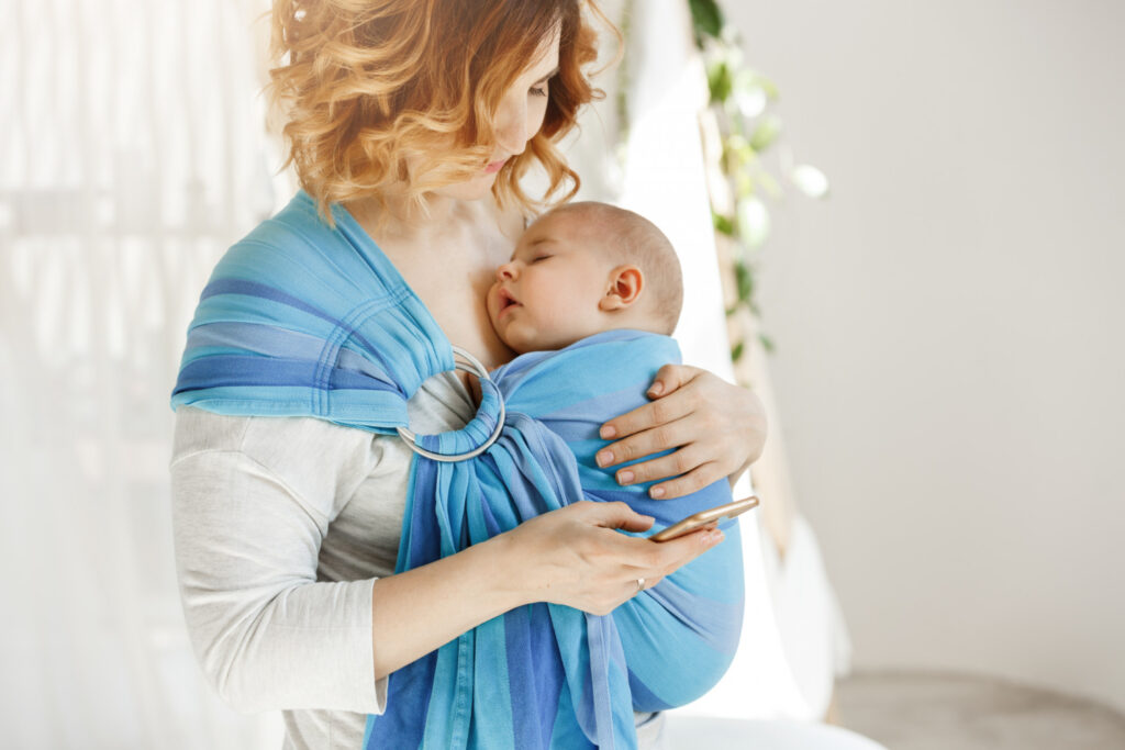 A woman with wavy hair holds a baby in a blue sling while using her smartphone, surrounded by soft, natural light.