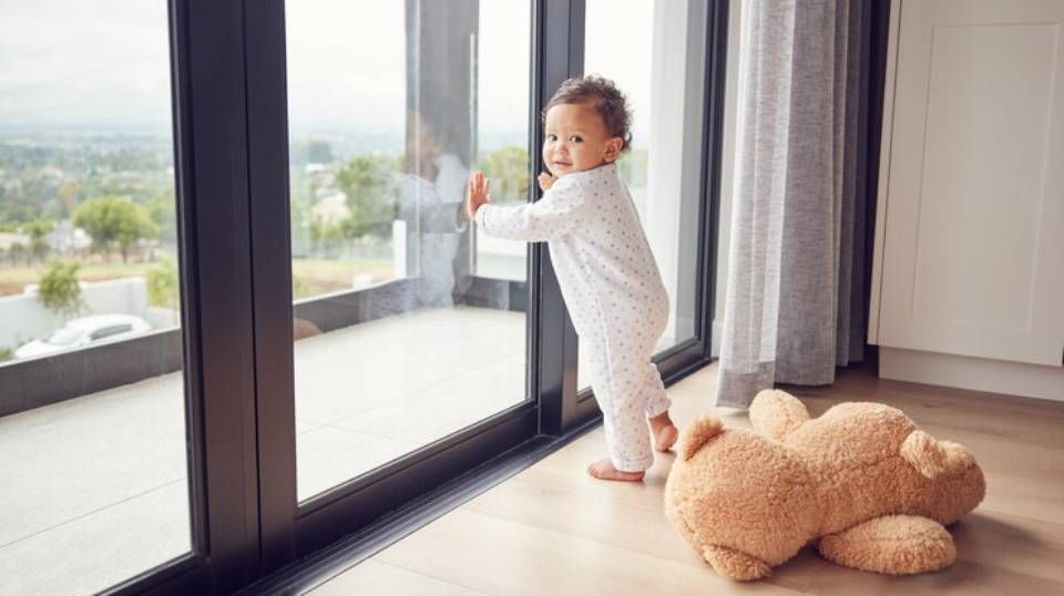 A toddler in polka-dot pajamas stands by a large glass door, gazing outside, while a plush teddy bear lies on the floor nearby.