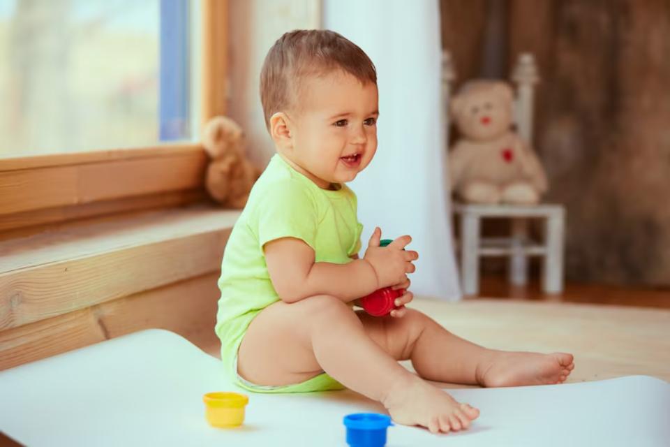  A toddler in a green outfit sits on a soft mat, holding colorful toys. A teddy bear is visible in a cozy room with warm lighting.