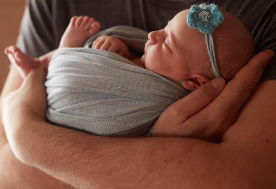  A caregiver gently holds a swaddled baby in a cozy setting, showcasing tiny feet and a blue hair accessory.
