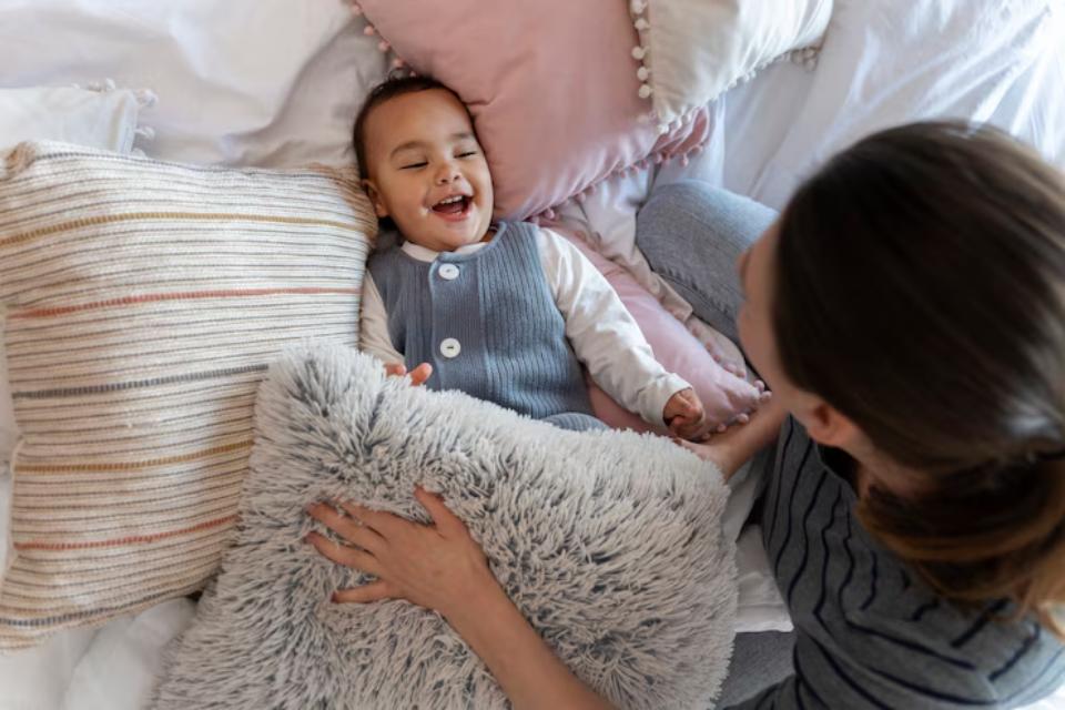  A close-up of a mother gently holding her baby's hand while they sit on a bed surrounded by soft pillows and blankets.