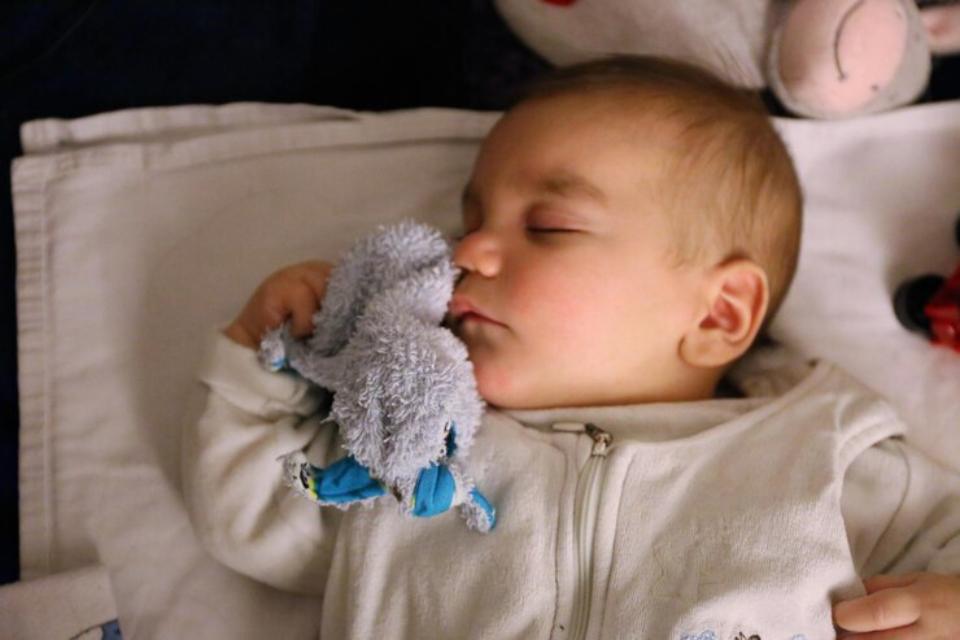  A small child lies on a white blanket, holding a soft stuffed toy while wearing a light zippered outfit, surrounded by colorful plush toys.