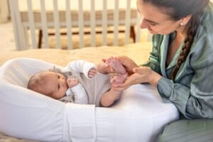 A caring adult gently holds a baby's feet, both seated on a cozy bed, with a crib visible in the background.