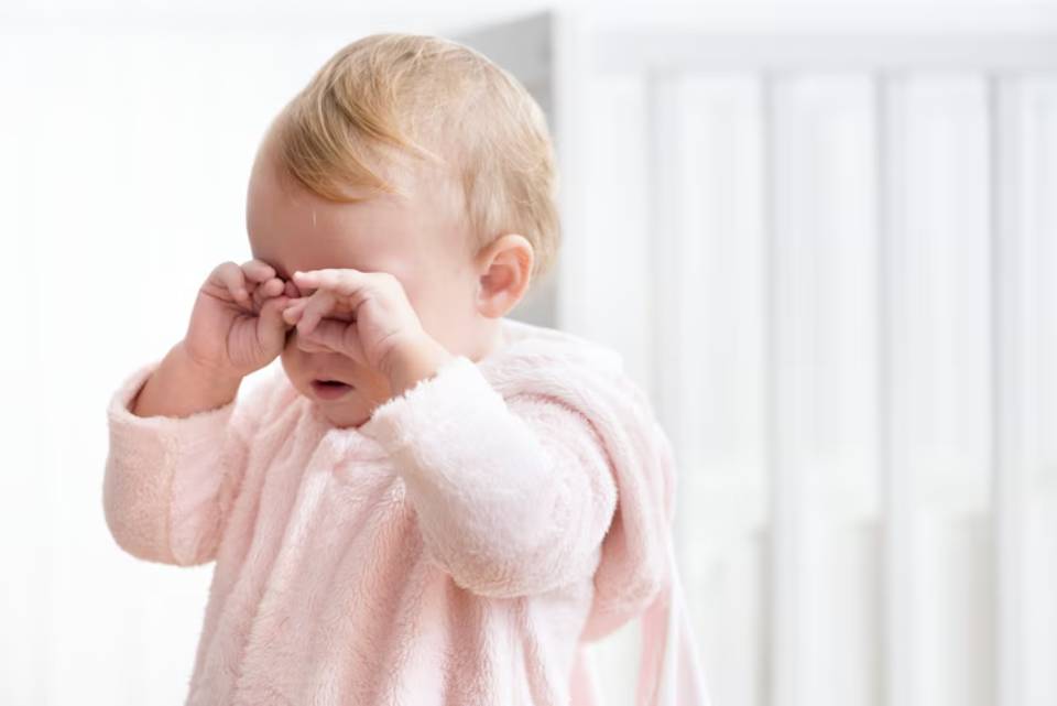 A toddler wearing a soft pink robe holds up their hands, surrounded by a bright, airy interior with soft-focus background elements.