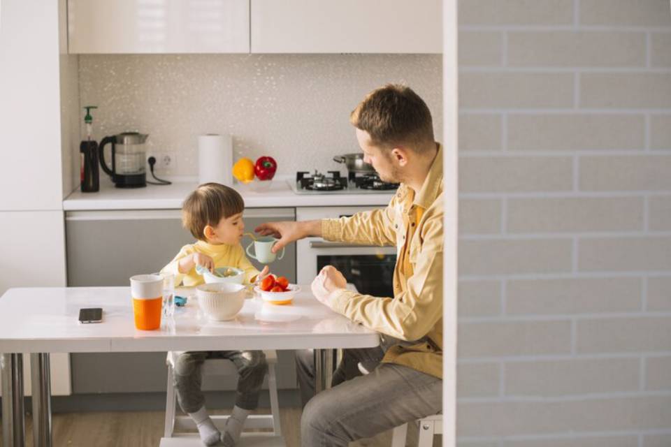 A child and an adult share a meal at a modern kitchen table, surrounded by bowls of food and colorful fruits.