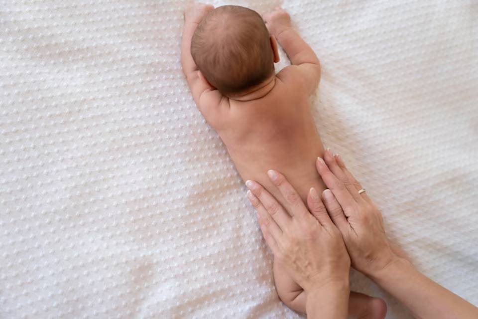 A close-up of a baby's bare back being gently massaged by two adult hands on a textured white blanket.