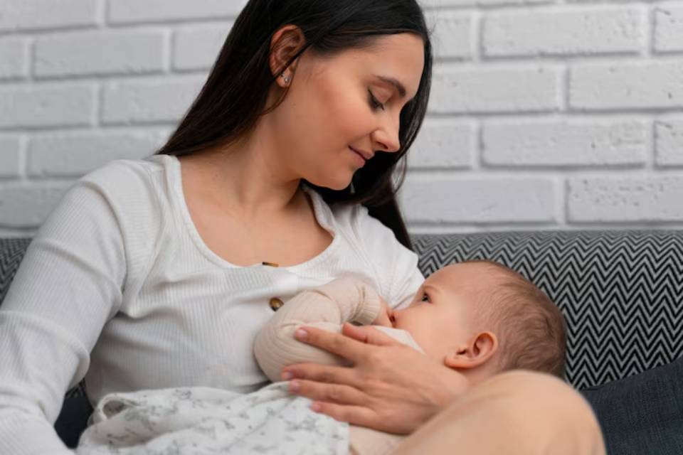 A woman in a white top cradles a baby in her arms while seated on a patterned couch against a white brick wall.