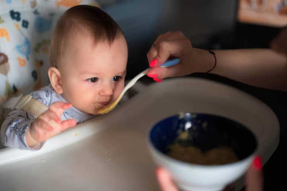 A caregiver feeds a baby from a spoon while a bowl of food sits on the table, highlighting a moment of mealtime interaction.