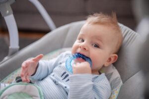 A cozy baby in a blue striped onesie sits in a gentle swing, reaching out curiously with one hand. Soft, blurred background ambiance.