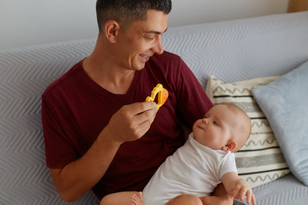 A parent sits on a couch, playfully showing a yellow fish toy to a baby cradled in their arm, surrounded by cozy cushions.