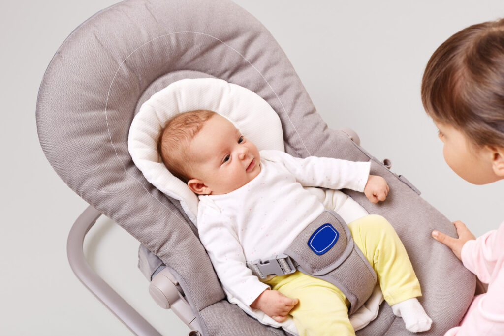 A baby in a soft, gray rocker chair wears a white long-sleeve outfit and yellow pants, with a harness securing them.