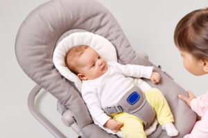 A baby in a soft, gray rocker chair wears a white long-sleeve outfit and yellow pants, with a harness securing them.