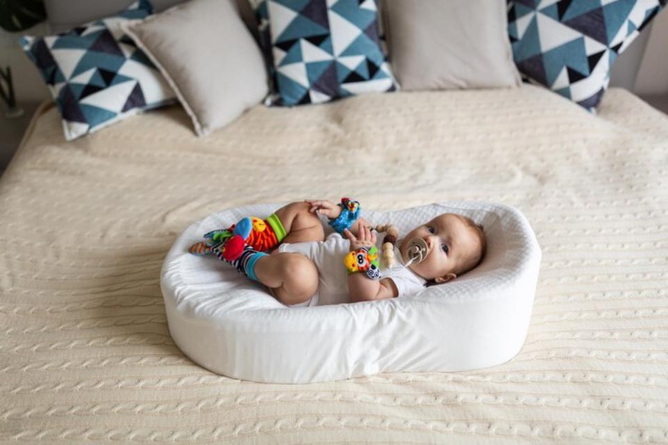  A playful toddler lies on a cream-colored bed in a fluffy white nest, surrounded by colorful toys and patterned pillows.
