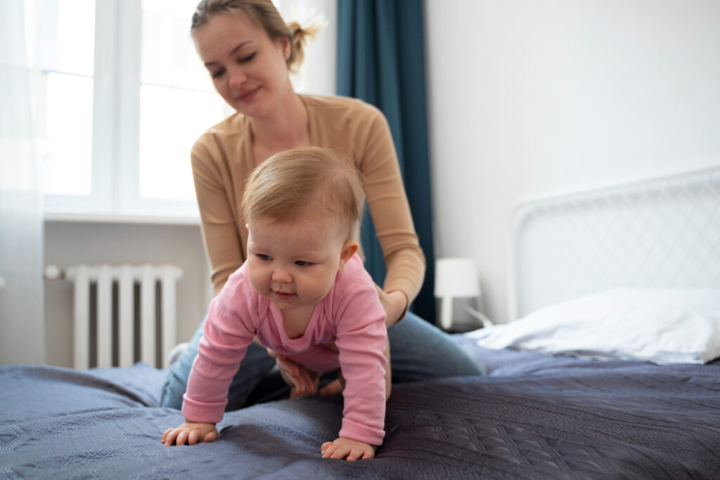 A baby in a pink outfit crawls on a bed, as an adult assists from behind in a bright, cozy room with natural light.