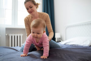 A baby in a pink outfit crawls on a bed, as an adult assists from behind in a bright, cozy room with natural light.
