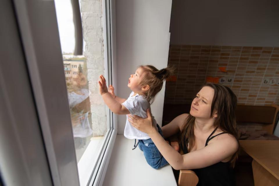  A toddler stands on a windowsill, reaching out toward the outside, while an adult supports them from behind. A cozy room is visible.