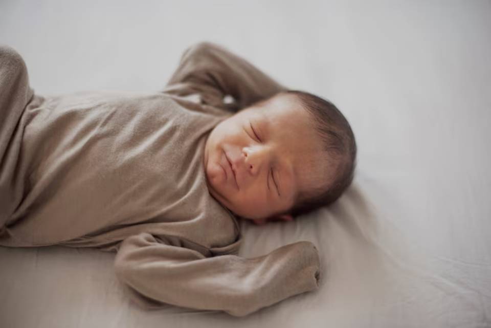 A peaceful newborn in a beige onesie, lying on a soft surface with arms raised, smiling slightly while asleep.