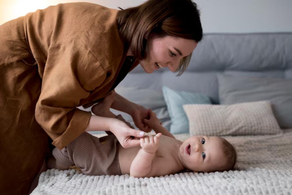  A caregiver gently changes an infant's diaper on a soft, textured blanket, surrounded by cozy pillows in a bright room.