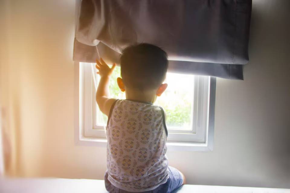 A young child in a patterned tank top sits on a bed, reaching toward a window partially covered by a gray curtain.