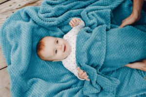 A baby wrapped in a soft, textured blue blanket, with playful hands reaching out, rests on a wooden surface.