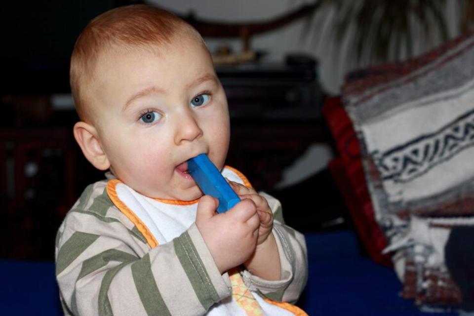  A baby in a striped shirt holds a small blue toy while sitting on a blue surface, with a patterned blanket in the background.