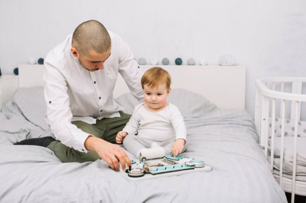 A parent and child sit on a bed, playing with a toy medical kit surrounded by soft bedding and a crib in a cozy, light-colored room.