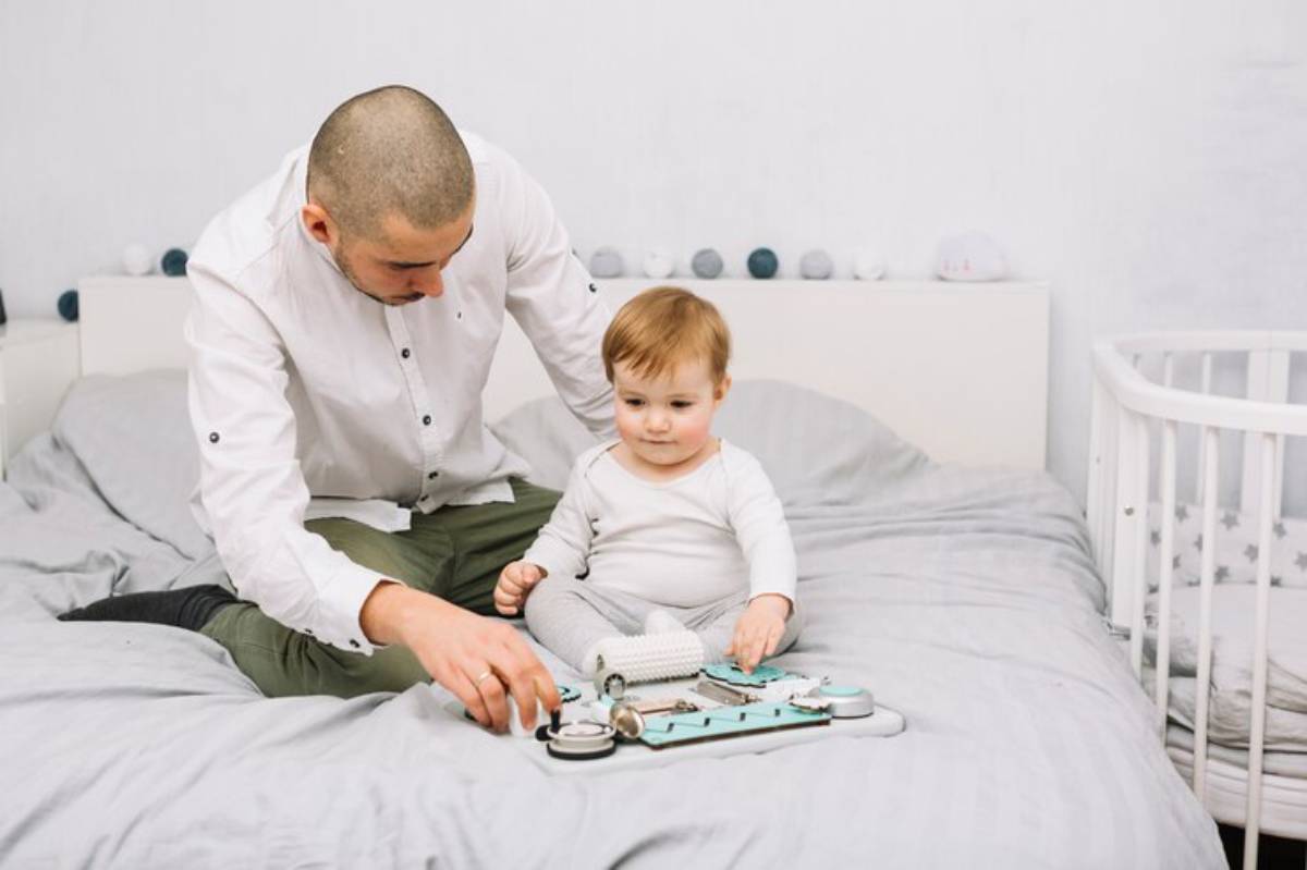 A parent and child sit on a bed, playing with a toy medical kit surrounded by soft bedding and a crib in a cozy, light-colored room.