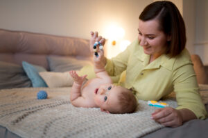 A parent plays with a baby on a cozy blanket, holding colorful toys, surrounded by soft pillows in a warmly lit room.