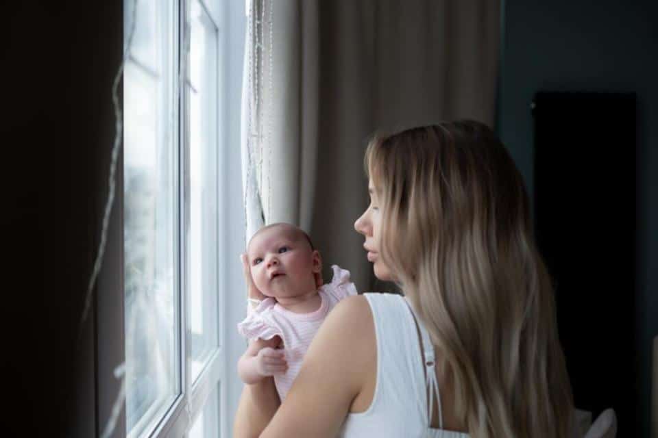 A woman holding a baby stands by a window, sunlight streaming in. Soft curtains frame the scene, creating a warm, cozy atmosphere.