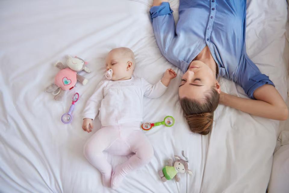  A mother and her baby lie on a white bed surrounded by colorful toys, enjoying a peaceful moment together.